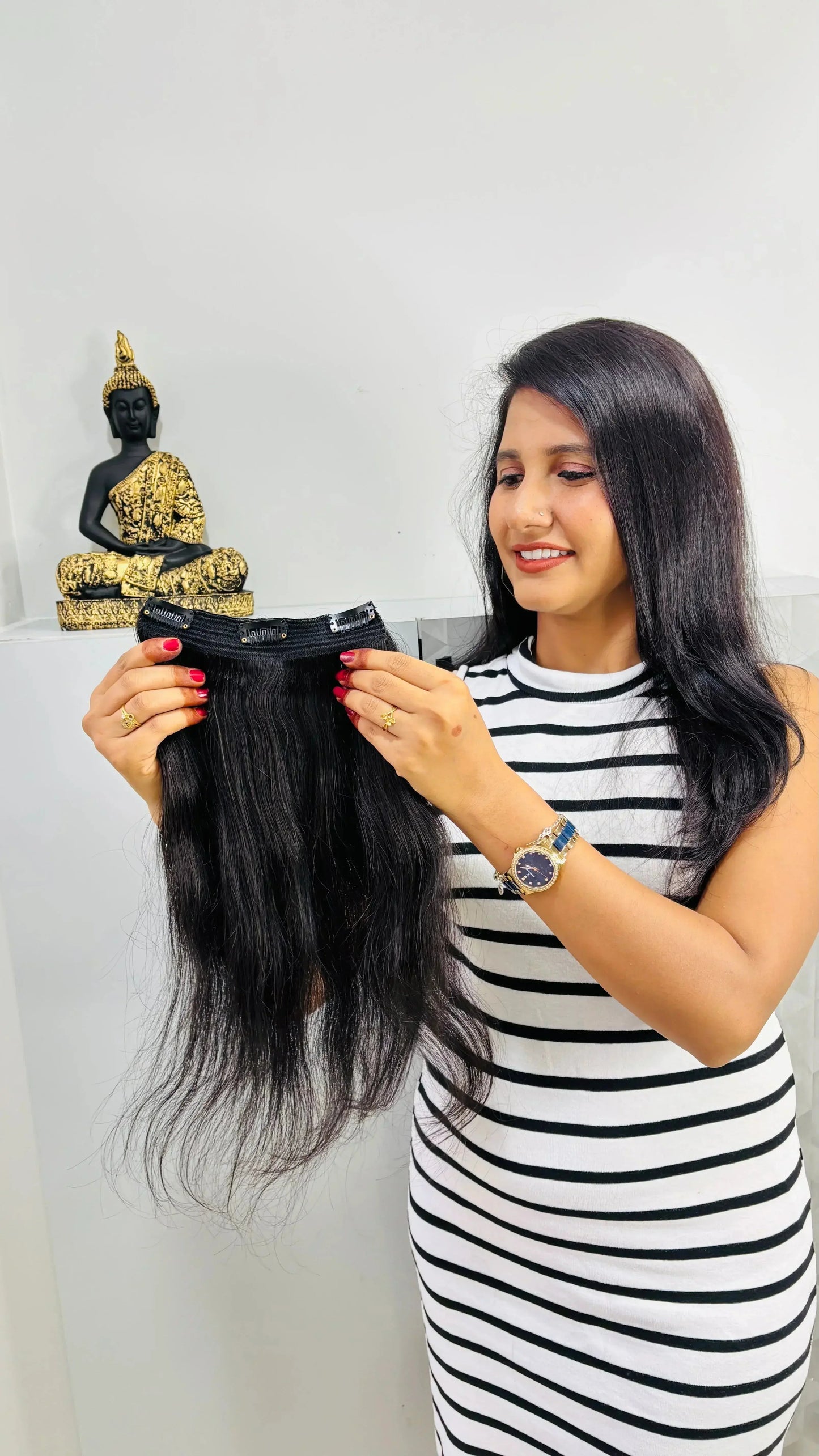 Woman in striped dress holding black clip-in hair extensions indoors with golden black Buddha statue in background