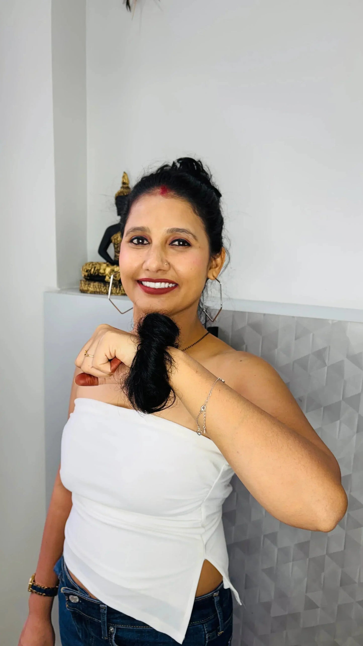 Smiling woman in white top and jeans holding a lock of her black hair indoors with kitchen decor