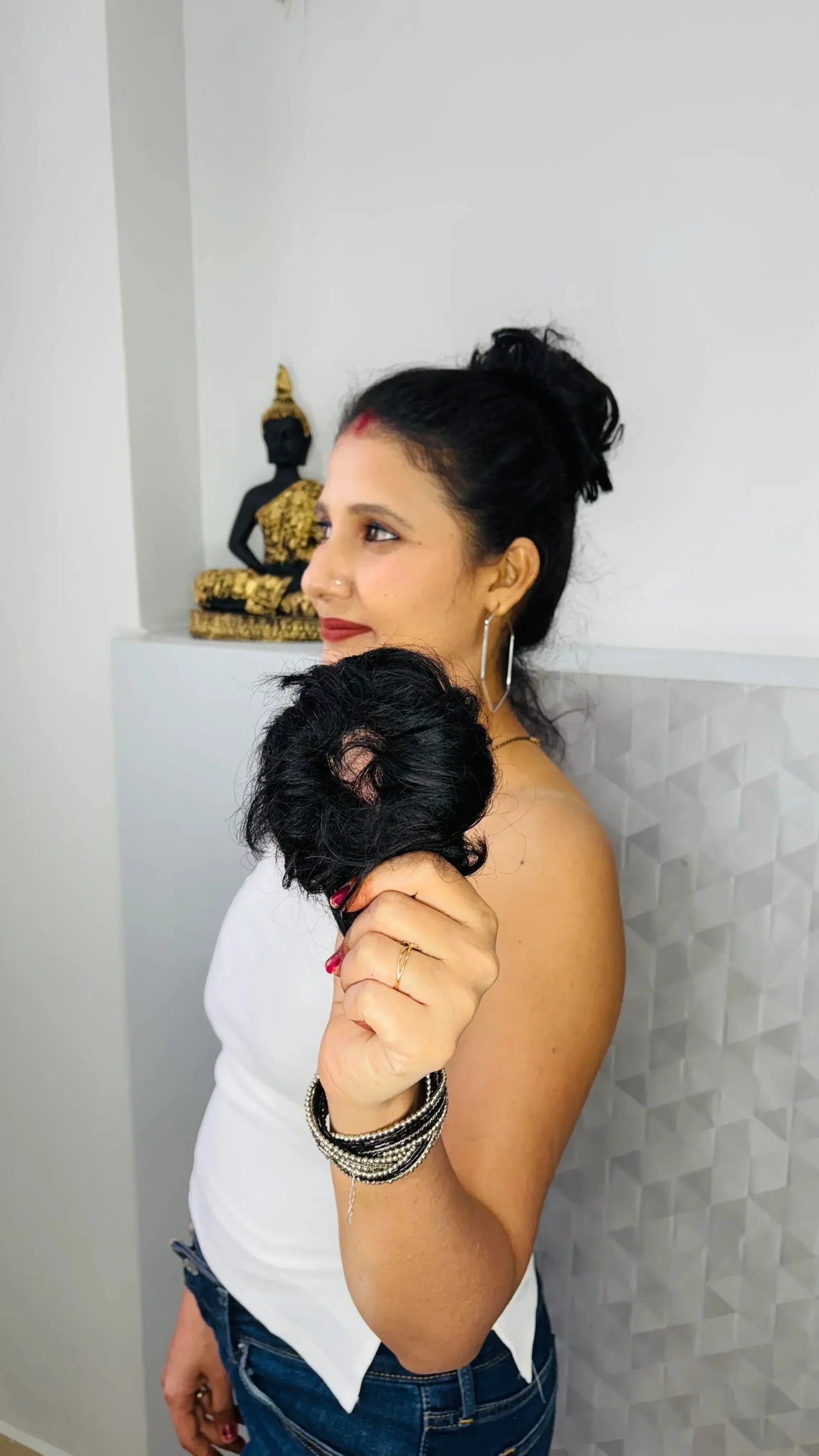 Woman with black hair bun holding black hair donut accessory wearing casual outfit with Buddha statue background