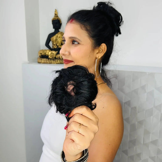 Woman holding a black hair bun extension, standing indoors with a Buddha statue in the background