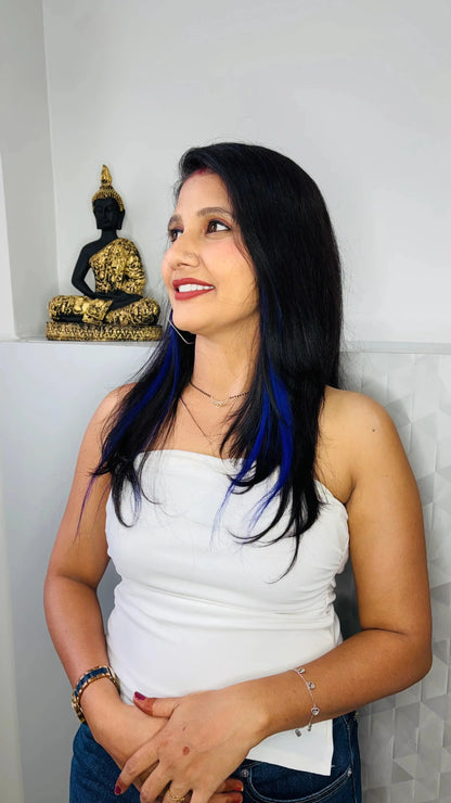 Woman with black hair and blue streaks wearing white strapless top and jewelry smiling near Buddha statue
