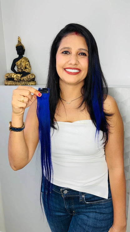 Smiling woman with black hair and blue highlights holding blue clip-in hair extension indoors with Buddha statue